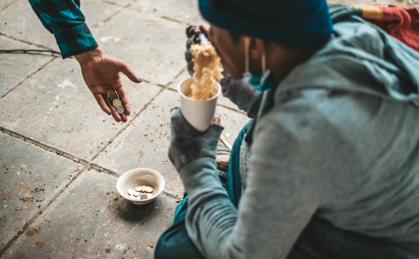 Foto ilustrativa de habitante de calle comiendo. Una mano le deposita monedas en un recipiente que tiene en el suelo. 